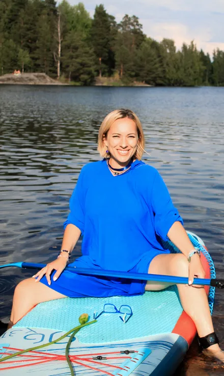 A beautiful blonde woman in a long blue dress sitting on a SUP board with the lake in the background.