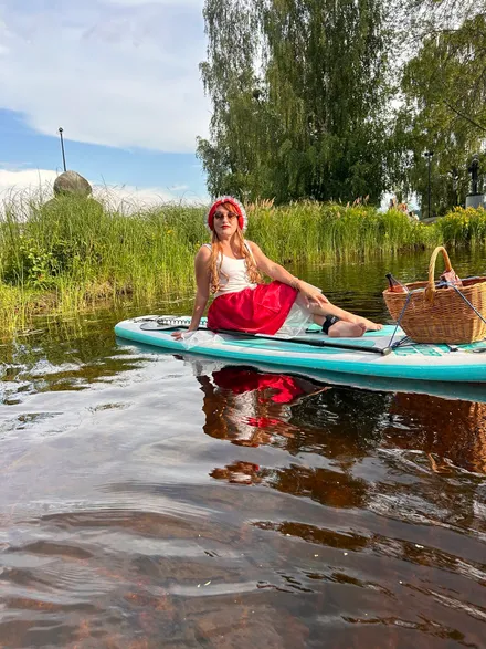 A participant in a bright Little Red Riding Hood costume sitting on a SUP board in a graceful pose, the board on the water, with grass, trees and sky in the background.