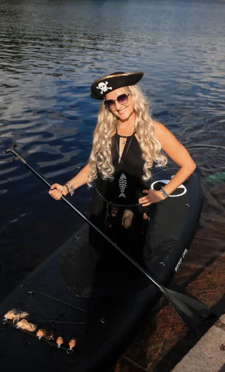 A blonde woman in a pirate costume standing on a SUP board on the water.