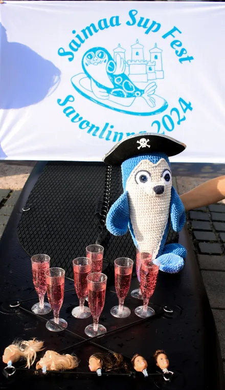 The knitted seal toy, the symbol of the festival, is standing on a SUP board wearing a pirate hat, with the festival flag in the background. In front of the seal are glasses with a drink.