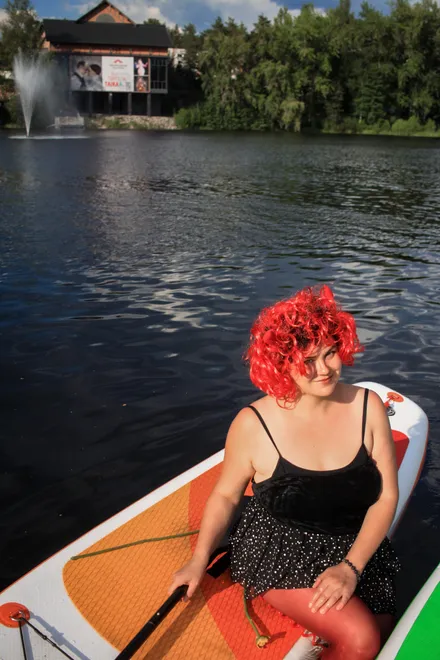 A participant in a bright red wig and a black tank‑dress sitting on a SUP board on the water, with the lake in the background.